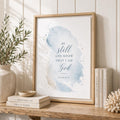 A framed wall art print leaning on a wooden shelf in a styled interior. The print features a soft blue watercolor wash with the text 'BE still AND KNOW THAT I AM God' and 'PSALM 46:10' in dark blue script.