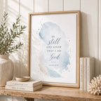 A framed wall art print leaning on a wooden shelf in a styled interior. The print features a soft blue watercolor wash with the text 'BE still AND KNOW THAT I AM God' and 'PSALM 46:10' in dark blue script.