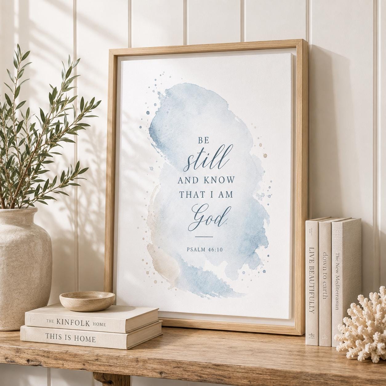 A framed wall art print leaning on a wooden shelf in a styled interior. The print features a soft blue watercolor wash with the text 'BE still AND KNOW THAT I AM God' and 'PSALM 46:10' in dark blue script.