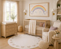 A beautifully styled nursery featuring a light wood crib with a cream throw blanket, a matching wood dresser, a round patterned rainbow rug, and a large framed rainbow artwork on the wall.
