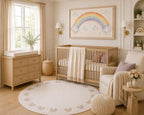 A beautifully styled nursery featuring a light wood crib with a cream throw blanket, a matching wood dresser, a round patterned rainbow rug, and a large framed rainbow artwork on the wall.