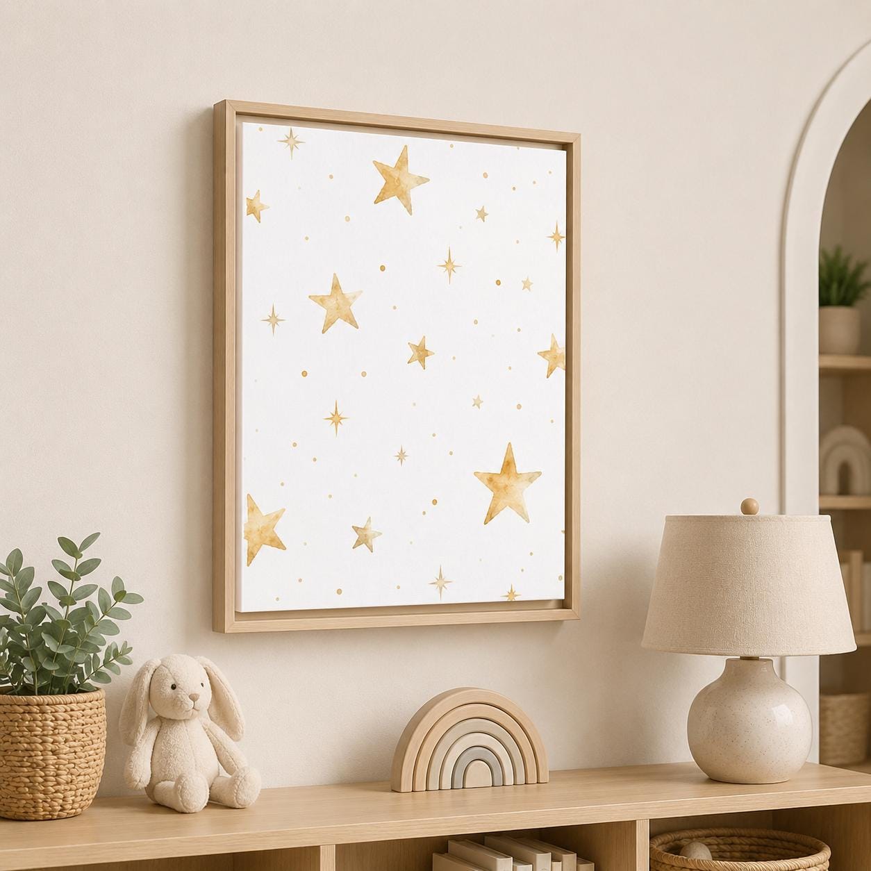 A framed wall art print featuring a white background with scattered gold watercolor stars, displayed in a styled nursery or children's room above a wooden shelf with a lamp, bunny toy, and plant.