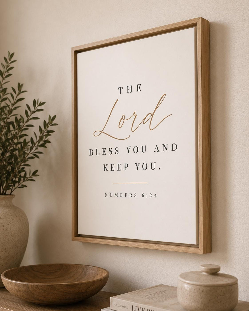Framed wall art with the Bible verse 'The Lord bless you and keep you. Numbers 6:24' in black serif text with 'The Lord' in gold script, displayed in a light wood frame on a beige wall above a wooden bowl and vase.