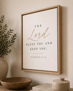 Framed wall art with the Bible verse 'The Lord bless you and keep you. Numbers 6:24' in black serif text with 'The Lord' in gold script, displayed in a light wood frame on a beige wall above a wooden bowl and vase.
