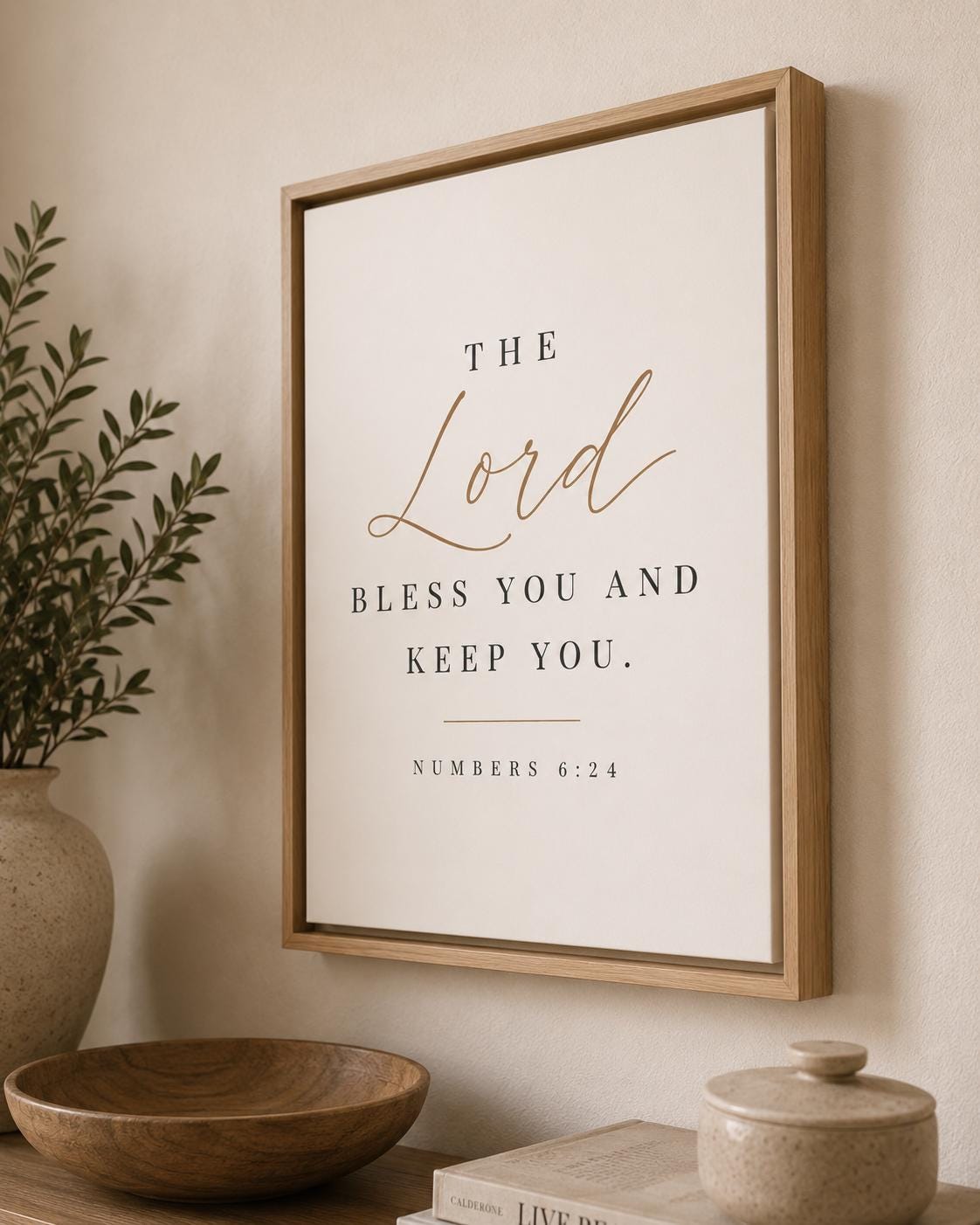 Framed wall art with the Bible verse 'The Lord bless you and keep you. Numbers 6:24' in black serif text with 'The Lord' in gold script, displayed in a light wood frame on a beige wall above a wooden bowl and vase.