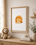 A framed watercolor art print of a sleeping golden lion displayed on a beige wall in a styled nursery or children's room. The light wood frame hangs above a wooden dresser decorated with a plush lion toy, stacked books, a small wooden rainbow, and a potted eucalyptus plant.