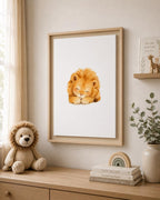A framed watercolor art print of a sleeping golden lion displayed on a beige wall in a styled nursery or children's room. The light wood frame hangs above a wooden dresser decorated with a plush lion toy, stacked books, a small wooden rainbow, and a potted eucalyptus plant.