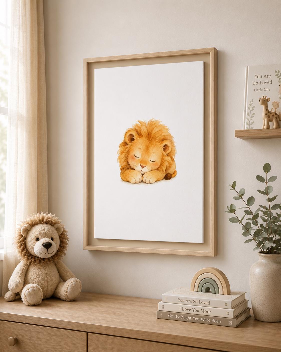 A framed watercolor art print of a sleeping golden lion displayed on a beige wall in a styled nursery or children's room. The light wood frame hangs above a wooden dresser decorated with a plush lion toy, stacked books, a small wooden rainbow, and a potted eucalyptus plant.