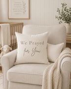 A cream-colored throw pillow with the text 'We Prayed for You' in brown lettering, styled on a light beige armchair in a nursery setting.