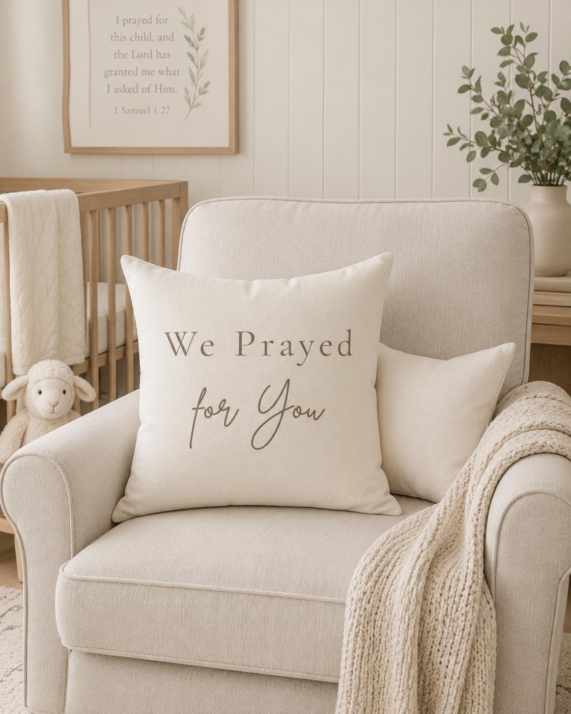 A cream-colored throw pillow with the text 'We Prayed for You' in brown lettering, styled on a light beige armchair in a nursery setting.