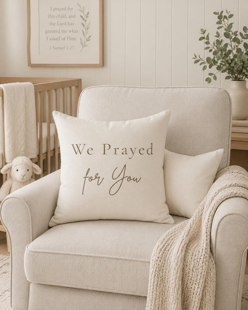 A cream-colored throw pillow with the text 'We Prayed for You' in brown lettering, styled on a light beige armchair in a nursery setting.