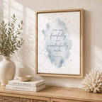 A framed wall art print with a watercolor splash and the quote 'YOU ARE fearfully and wonderfully MADE. PSALM 139:4' hangs on a beige wall above a wooden console table styled with books, a vase, and coral.