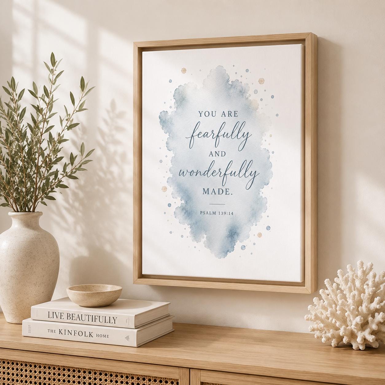 A framed wall art print with a watercolor splash and the quote 'YOU ARE fearfully and wonderfully MADE. PSALM 139:4' hangs on a beige wall above a wooden console table styled with books, a vase, and coral.