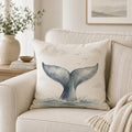 A square throw pillow with a watercolor whale tail design in blue-gray tones, resting on a light beige sofa in a styled living room setting.