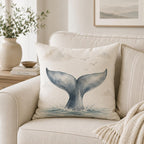 A square throw pillow with a watercolor whale tail design in blue-gray tones, resting on a light beige sofa in a styled living room setting.