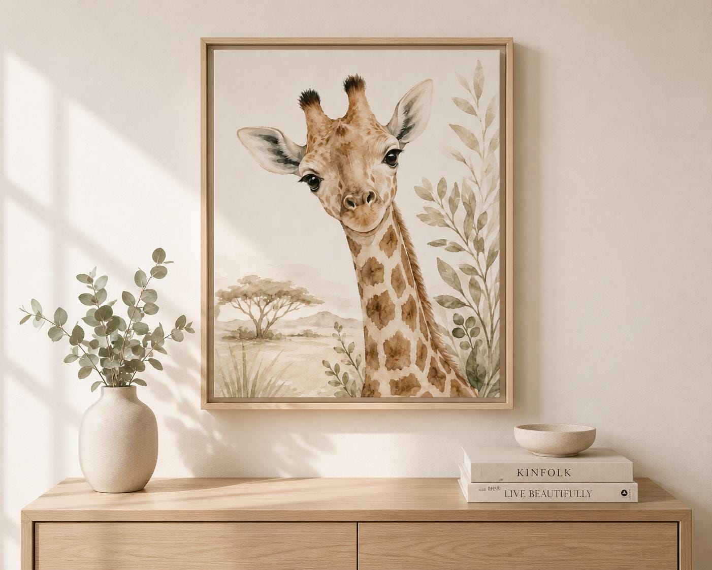 A framed watercolor-style giraffe wall art print hangs on a beige wall above a light wood console table. The scene includes a white vase with greenery on the left and a stack of books with a small bowl on the right.