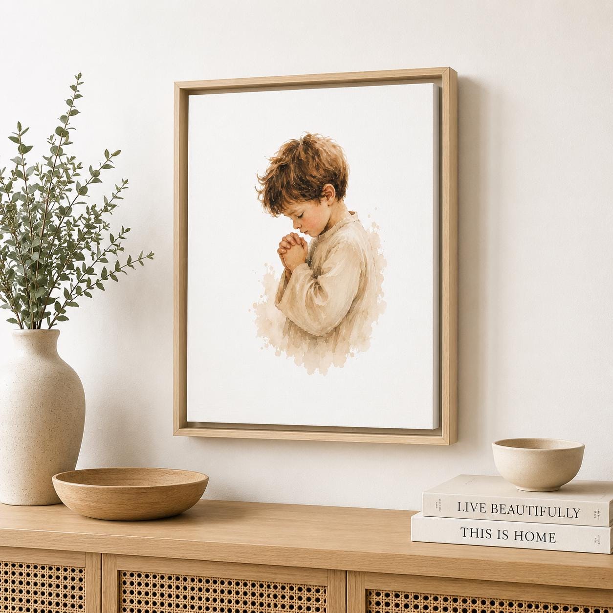 A framed art print featuring a watercolor-style illustration of a young boy in a beige robe, displayed on a light wood console table against a white wall, styled with a vase of greenery, a bowl, and books.
