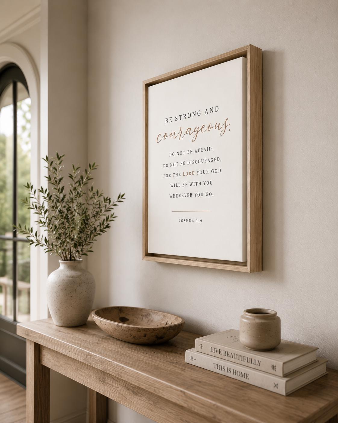 A framed wall art print with a Bible verse hangs on a beige wall above a wooden console table styled with a vase of greenery, a bowl, and stacked books.