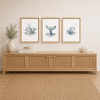 Three framed whale art prints in light wood frames hang above a light wood sideboard with woven cabinet panels, styled in a bright, neutral room with a vase of greenery and a woven rug.