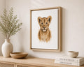 A framed watercolor-style print of a lion cub hanging on a light beige wall above a wooden console table. The artwork shows a young lion cub centered on a white background, enclosed in a light wood frame.