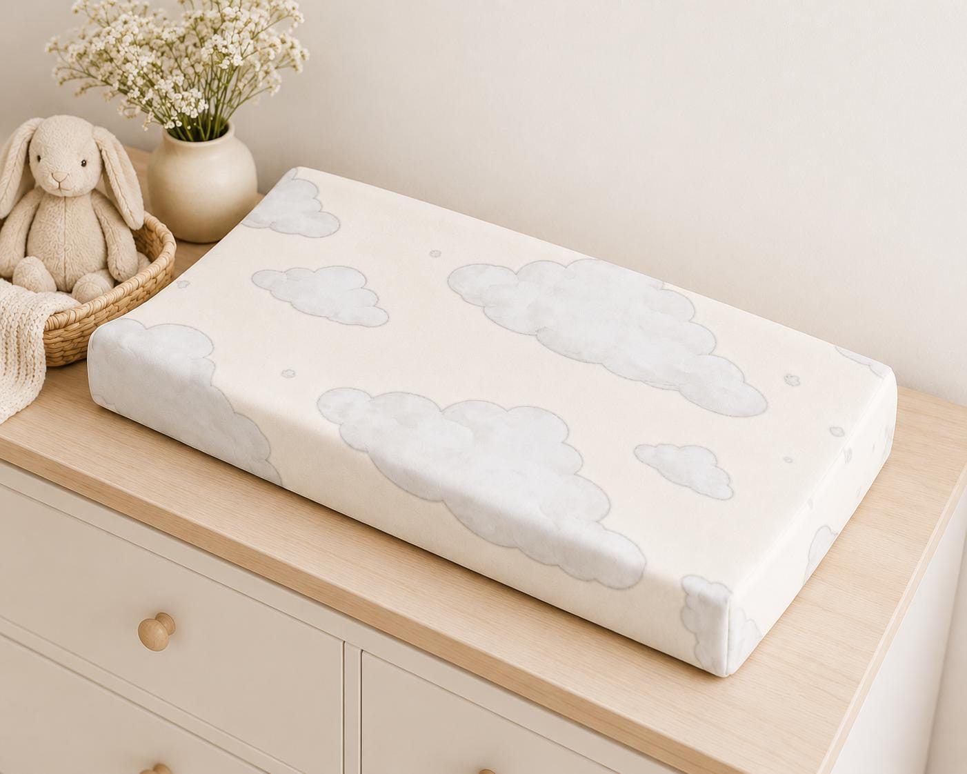 A rectangular baby changing pad with a cream background and light gray cloud pattern, resting on a light wood dresser in a nursery setting next to a plush bunny and a vase of small white flowers.