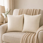 Two square cream throw pillows on a beige upholstered sofa with a chunky knit beige throw blanket draped over the chair in a softly lit living room.