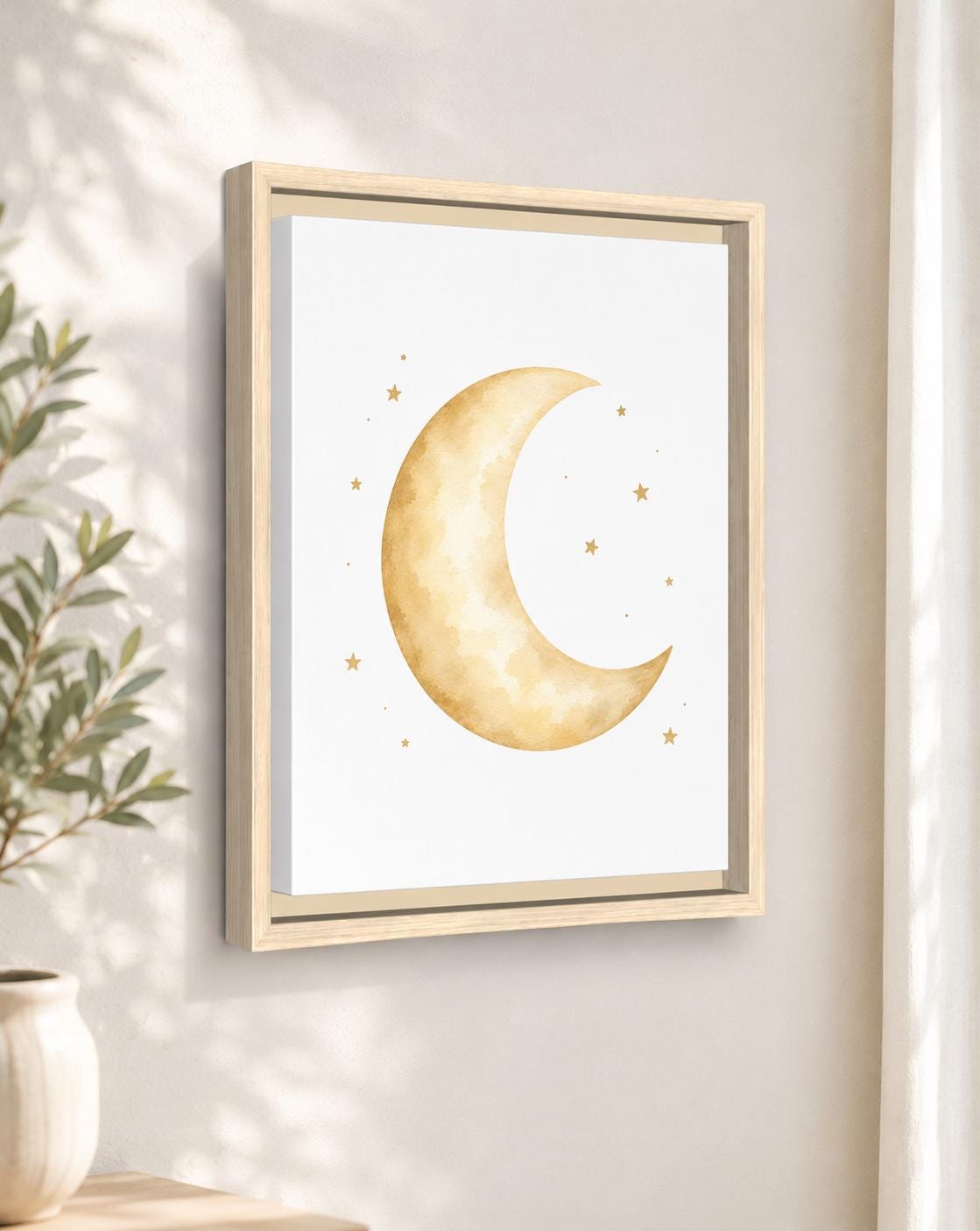 Framed wall art featuring a soft watercolor crescent moon and small stars in warm gold and beige tones on a white background, shown hanging on a light neutral wall beside a plant.