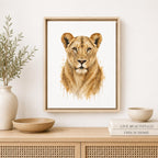 A framed watercolor art print of a lioness face, displayed on a light wood console table in a styled room setting with a vase, bowls, and books.