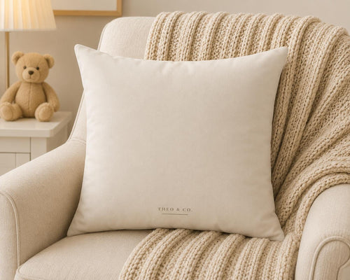 A square off-white throw pillow with 'THEO & CO.' printed in a subtle gold font at the bottom, styled on a cream armchair with a chunky knit beige blanket.