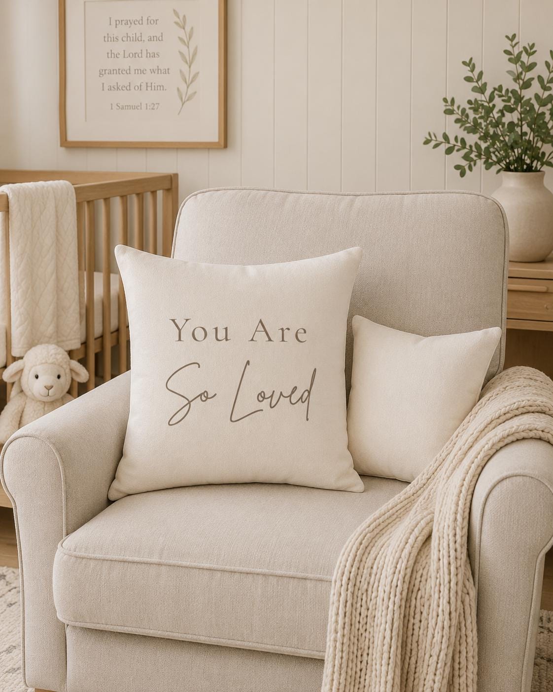 A cream-colored throw pillow with the text 'You Are So Loved' in dark gray lettering, styled on a light beige armchair in a nursery setting.