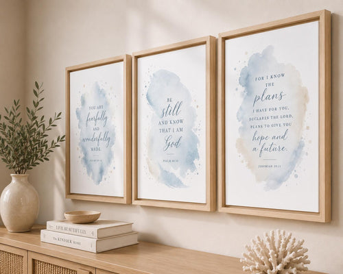 Three framed watercolor scripture prints with light blue and beige washes hang on a beige wall above a wooden console table with books, a vase, and a coral decor piece.
