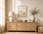 A light natural wood dresser in a styled nursery or bedroom setting, decorated with a table lamp, a framed art print of foxes, a woven basket, books, and a vase with greenery.