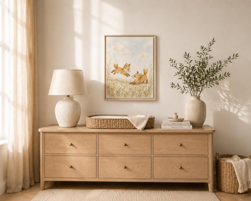 A light natural wood dresser in a styled nursery or bedroom setting, decorated with a table lamp, a framed art print of foxes, a woven basket, books, and a vase with greenery.