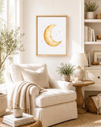 A framed art print featuring a gold watercolor crescent moon and small stars on a white background, styled in a warm, neutral-toned living room above a cream armchair.