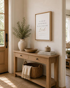 A framed wall art print with the Bible verse 'AS FOR ME AND MY HOUSE, WE WILL SERVE THE Lord' displayed in a light wood frame on a beige wall above a wooden console table in a bright, modern entryway.