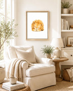 A framed watercolor art print of a sleeping golden lion hanging on a cream wall in a beautifully styled living room with a beige armchair, lamp, and books.