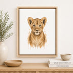Framed watercolor art print of a lion cub centered on a light wall above a wooden console, with a vase, bowls, and stacked books in a styled interior setting.