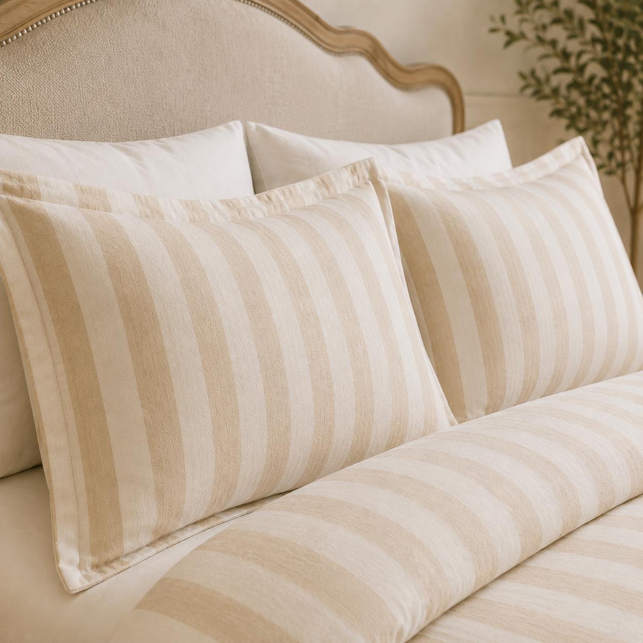 Beige and cream striped pillowcases and matching striped bedding on a bed with a light upholstered headboard.