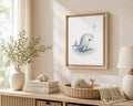 A framed whale art print hangs on a beige wall above a light wood console table styled with a vase of greenery, stacked books, a woven basket, and a lamp in a softly lit room.