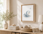A framed whale art print hangs on a beige wall above a light wood console table styled with a vase of greenery, stacked books, a woven basket, and a lamp in a softly lit room.