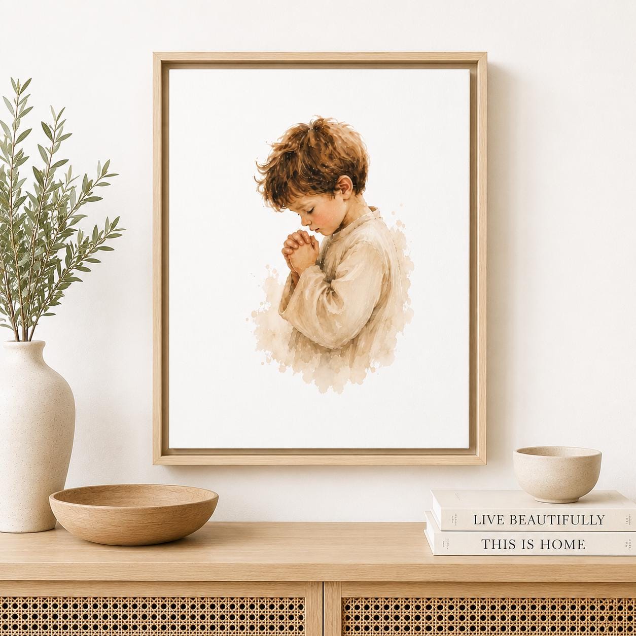 A framed art print featuring a watercolor-style illustration of a young boy in a beige robe, shown hanging on a white wall above a wooden console table with a vase, bowl, and books.