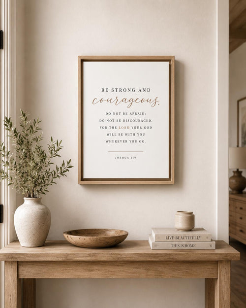 A framed inspirational Bible verse Joshua 1:9 print hangs on a beige wall above a wooden console table styled with a vase of greenery, a bowl, stacked books, and a small candle.