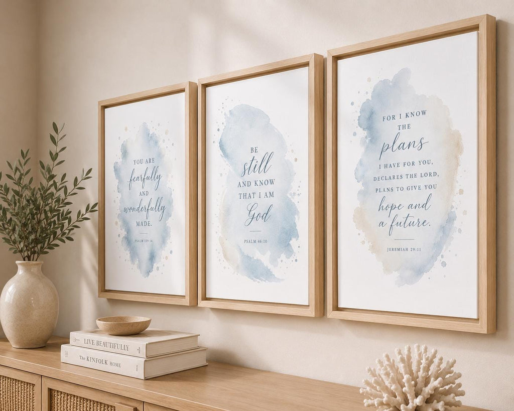 Three framed watercolor art prints with Bible verses hang on a beige wall above a wooden console table. The prints feature soft blue and beige watercolor washes with elegant script typography.