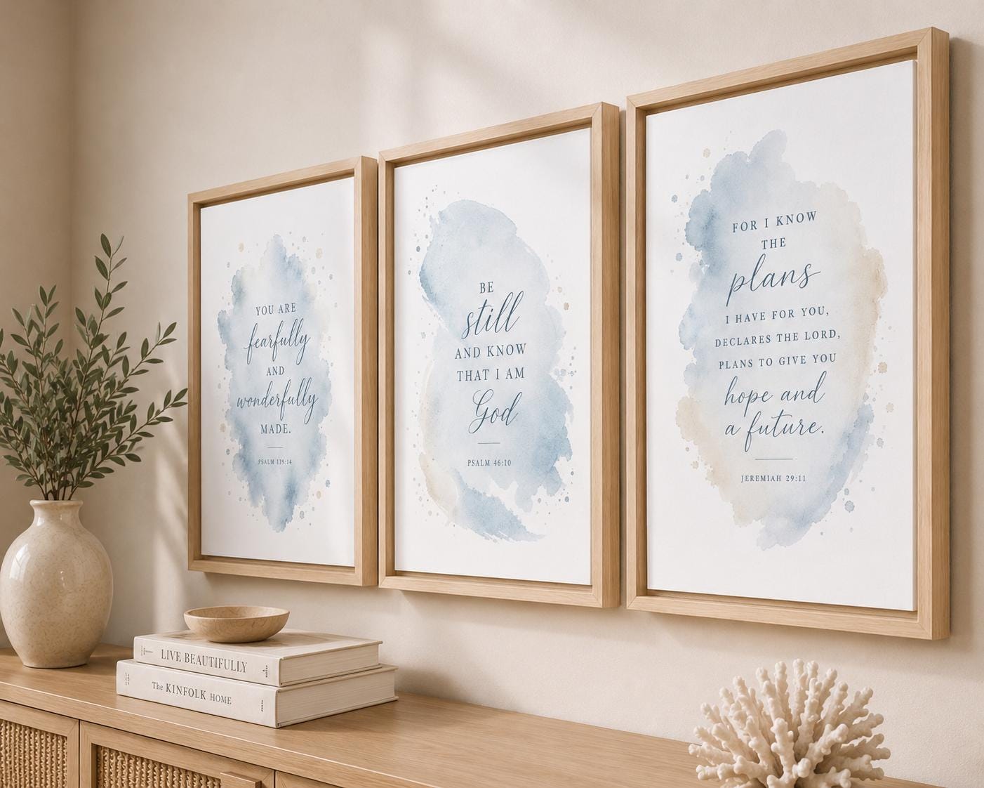 Three framed watercolor art prints with Bible verses hang on a beige wall above a wooden console table. The prints feature soft blue and beige watercolor washes with elegant script typography.