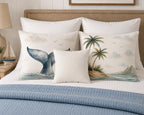 A styled bed with white bedding and a blue ribbed throw, featuring decorative throw pillows. Two larger pillows have a cream background with a blue whale tail and a tropical palm island scene, while a smaller square pillow in front is solid cream.