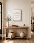 A framed wall art print with a quote hangs on a beige wall above a wooden console table in a styled home interior. The print has a light wood frame and black typography on a warm off-white background.