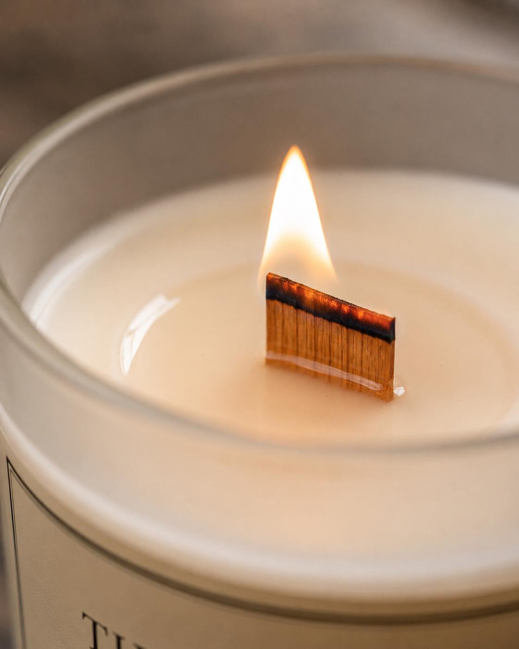 A close-up of a lit candle in a frosted glass jar, with a match-shaped wick burning in the center of the creamy off-white wax.