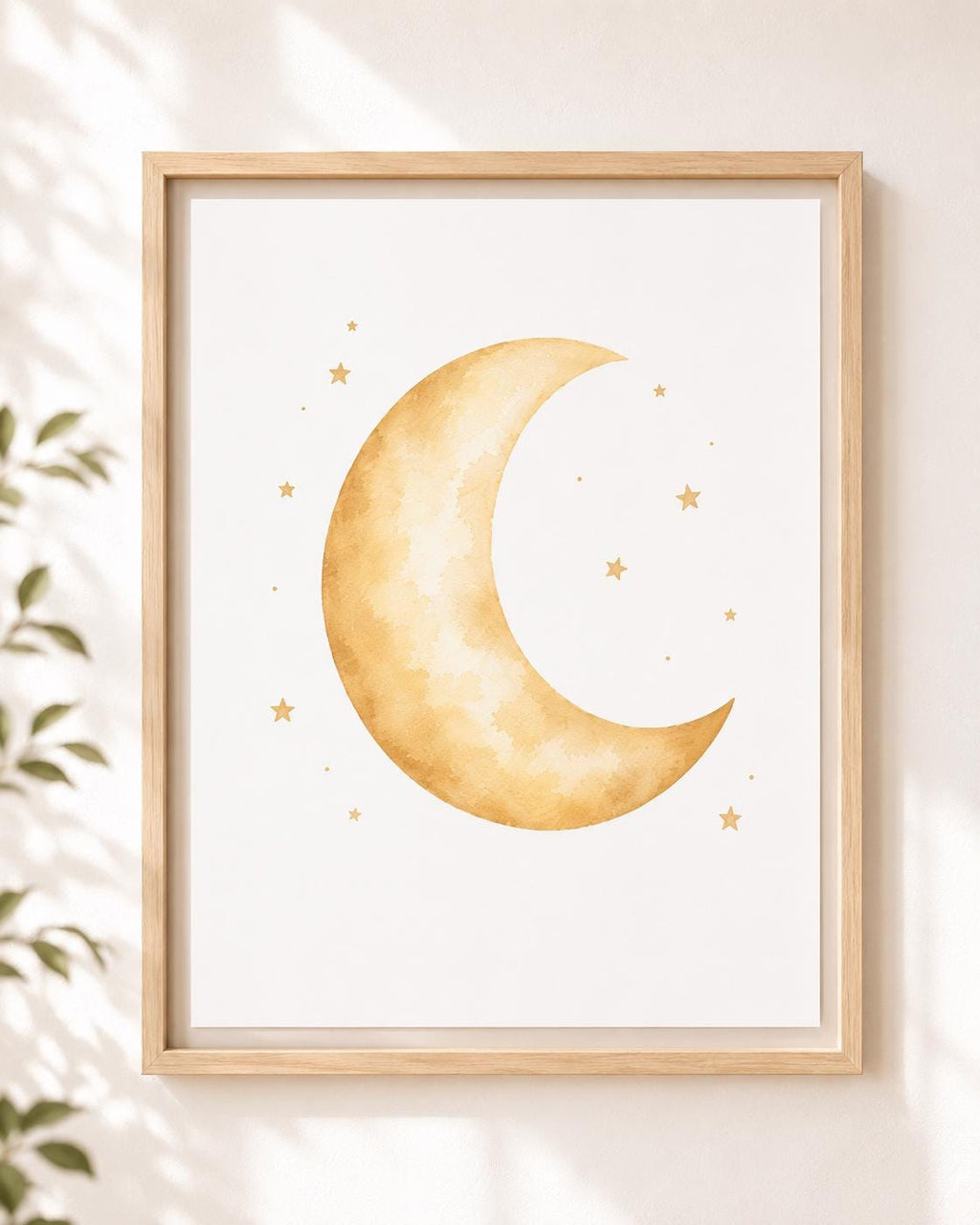 Framed wall art featuring a watercolor-style gold crescent moon and small stars on a white background, shown hanging on a light wall with soft shadows and a plant leaf at the side.