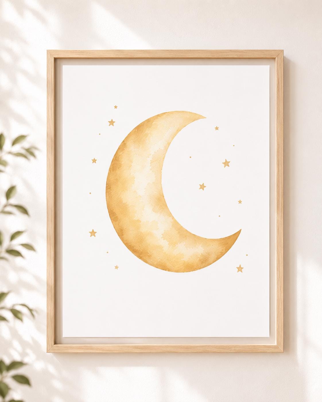 Framed wall art featuring a watercolor-style gold crescent moon and small stars on a white background, shown hanging on a light wall with soft shadows and a plant leaf at the side.