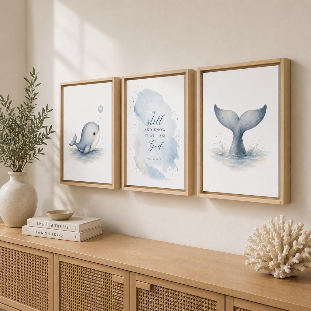 Three framed wall art prints hang above a wooden console table in a styled interior. The prints feature watercolor whale and mermaid tail illustrations with soft blue, gray, and white tones.